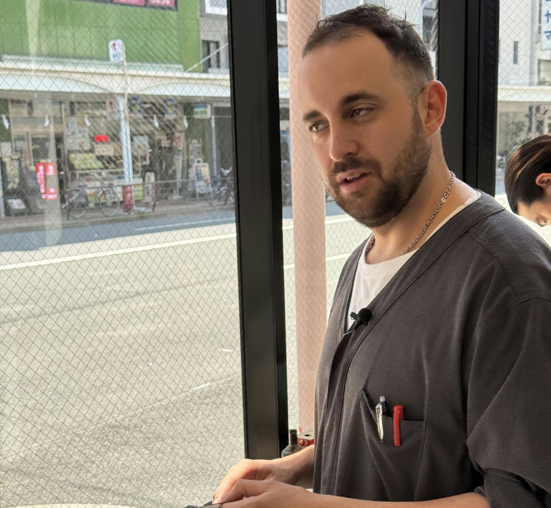 A man in a dark gray chef's coat demonstrates proper knife sharpening technique using a traditional whetstone. He's standing in front of a window that fills the entire frame.